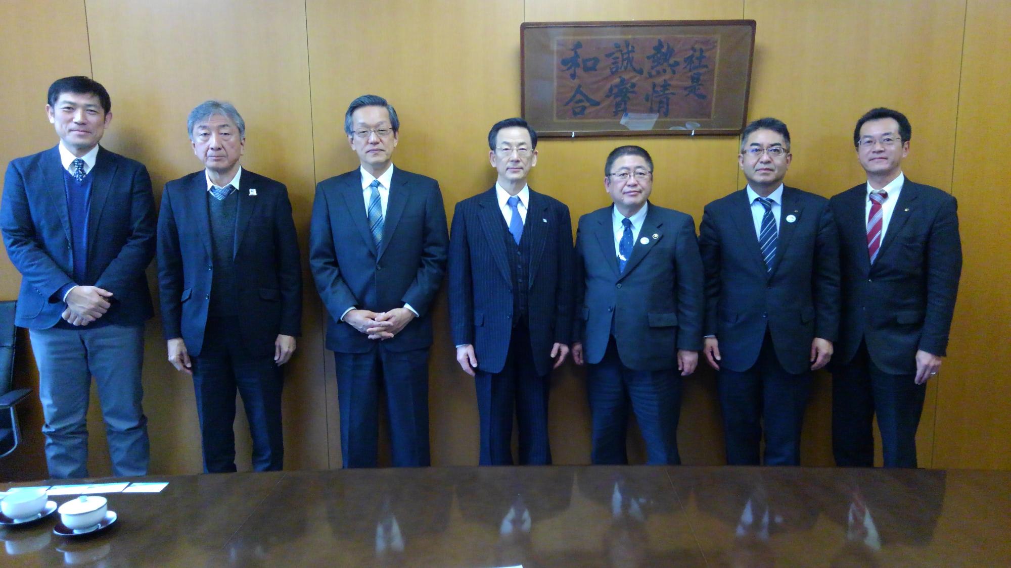 壁の前で並んで立つ市長と6名の男性の記念写真