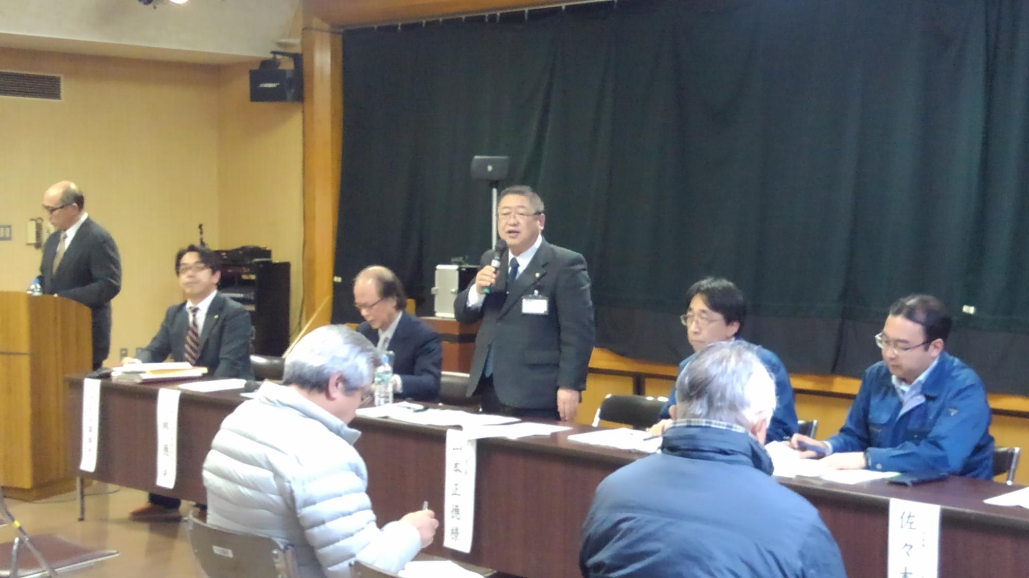 名前札が垂らされたテーブルの席の中央で、右手にマイクを持ち立って話している様子の市長の写真