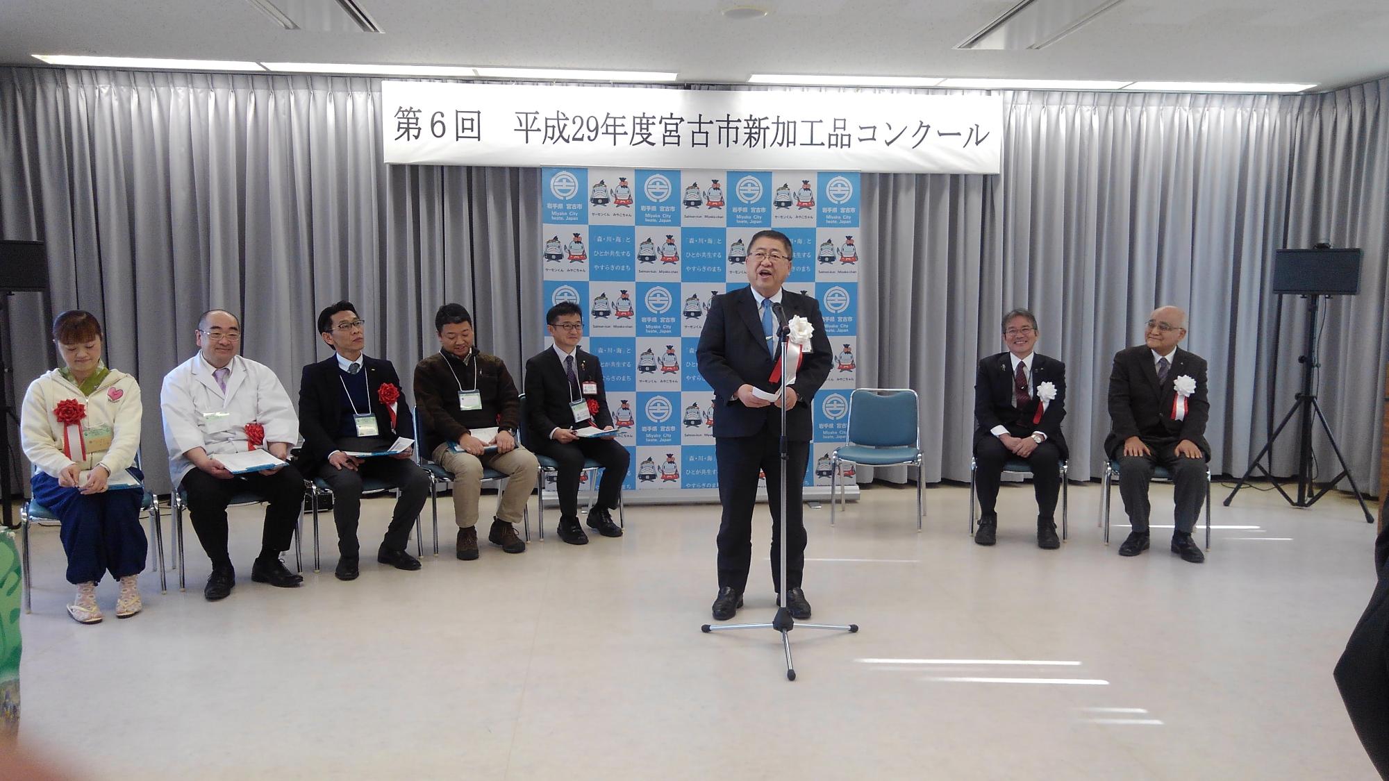 「第6回平成29年度宮古市新加工品コンクール」と書かれた横断幕が吊るされた会場で、スタンドマイクに向かい話をする様子の市長と、後方で並べられた椅子に座る7名の関係者の写真