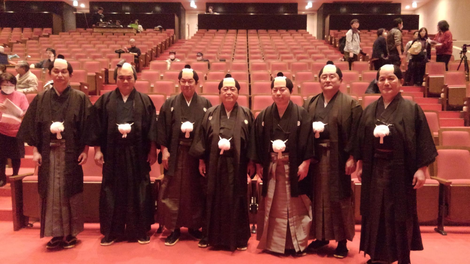 会場の椅子の前で、袴を着てちょんまげのかつらをかぶった市長と6名の男性が横一列に並んでいる記念写真