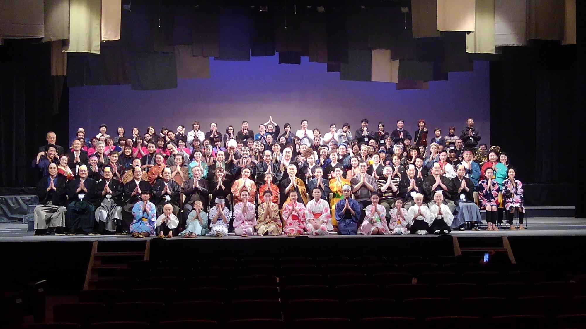 舞台上で着物や袴など演劇の衣装を着た大勢の演者の方々が集合している記念写真