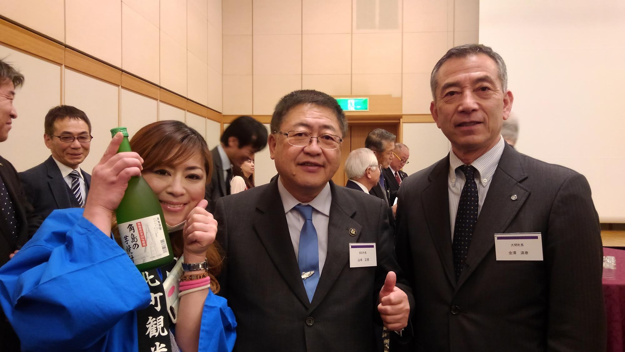 お酒の瓶を持ち青い法被を着た女性と市長、男性が並んで立つ記念写真