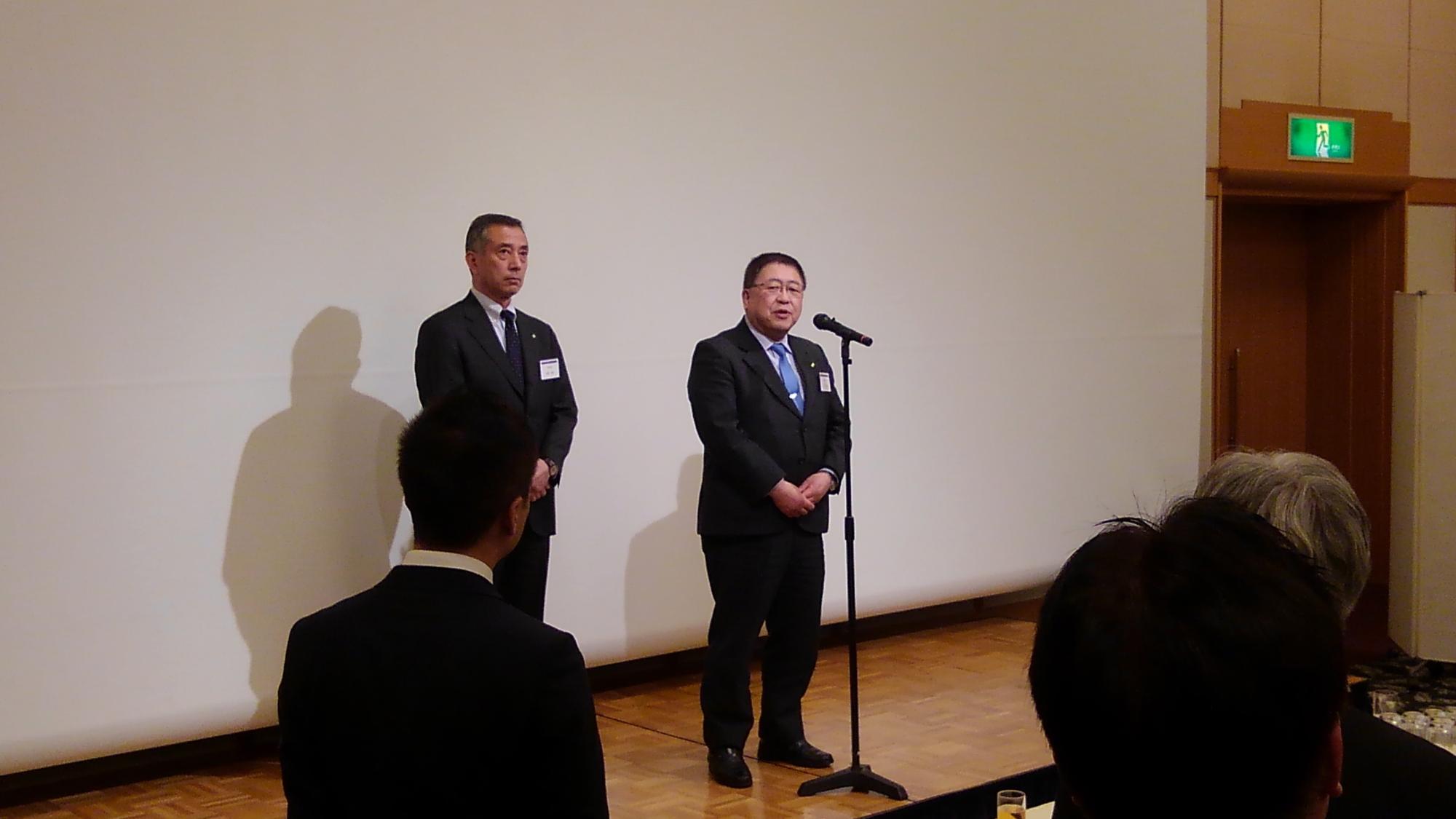 スクリーンの前でスタンドマイクに向かい話をする様子の市長と、斜め後ろに立つ男性の写真