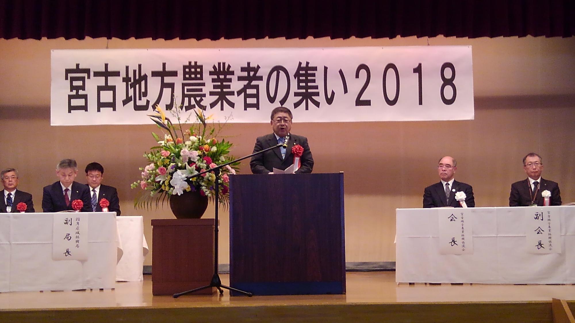 「宮古地方農業者の集い2018」と書かれた横断幕が吊るされた舞台上で、演台の前に置かれたスタンドマイクに向かい話をする様子の市長の写真
