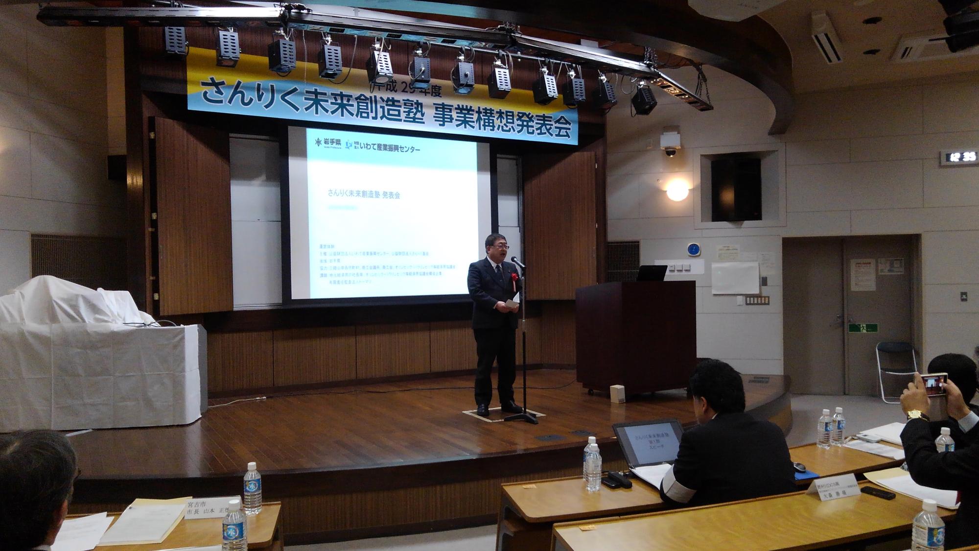 「さんりく未来創造塾 事業構想発表会」と書かれた横断幕が吊るされ、画面が映されたスクリーンが設置された舞台上で、スタンドマイクに向かい話をする様子の市長の写真