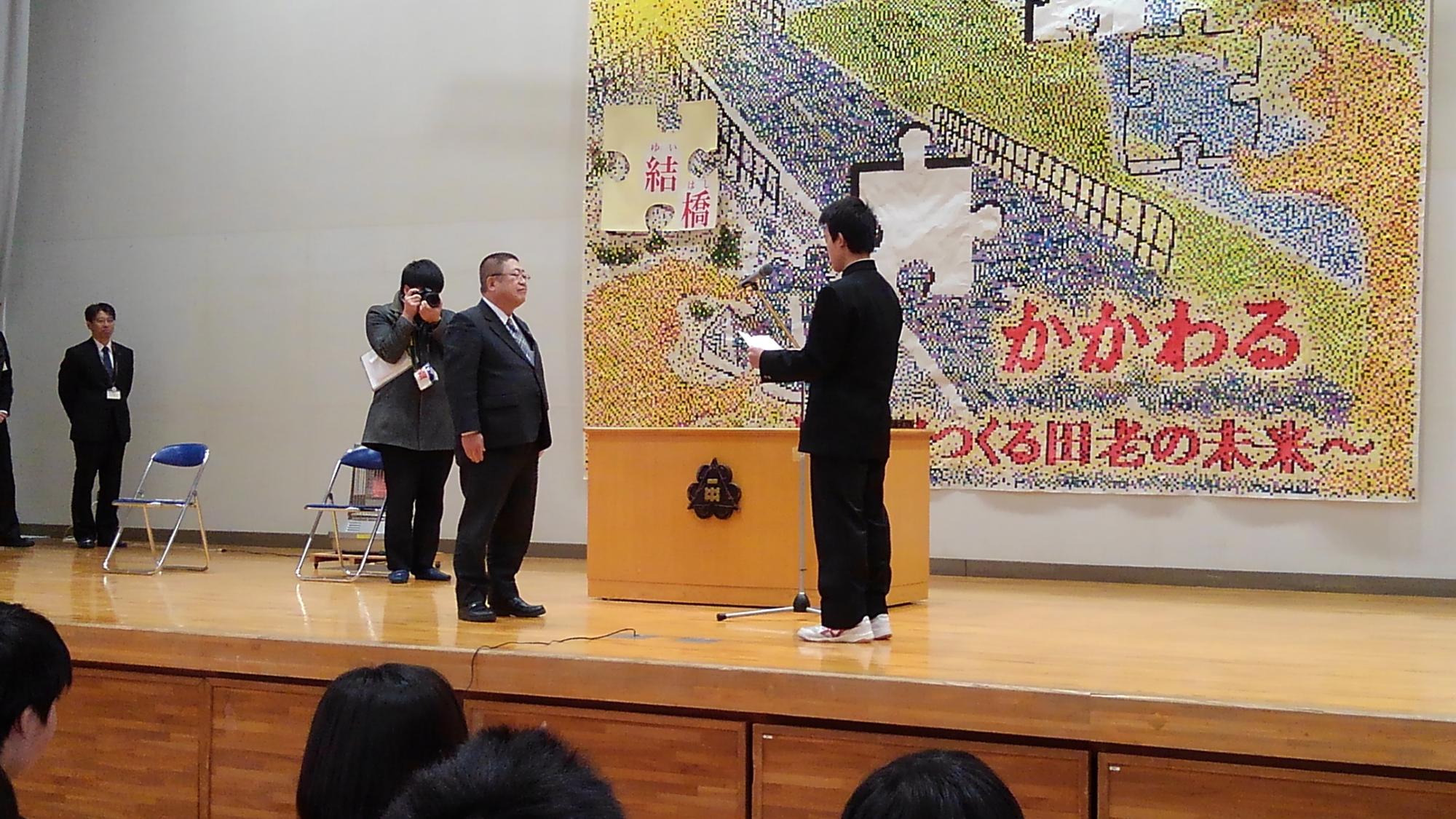 舞台上で、紙を広げて読んでいる様子の制服を着た男子中学生と向かい合う市長を写した写真