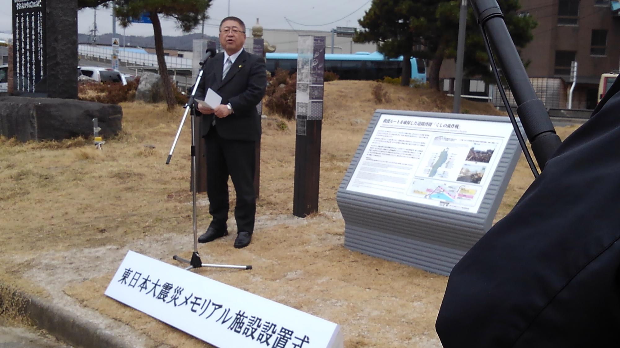 写真や文字が書かれた看板の斜め前で、スタンドマイクに向かい話をする様子の市長の写真