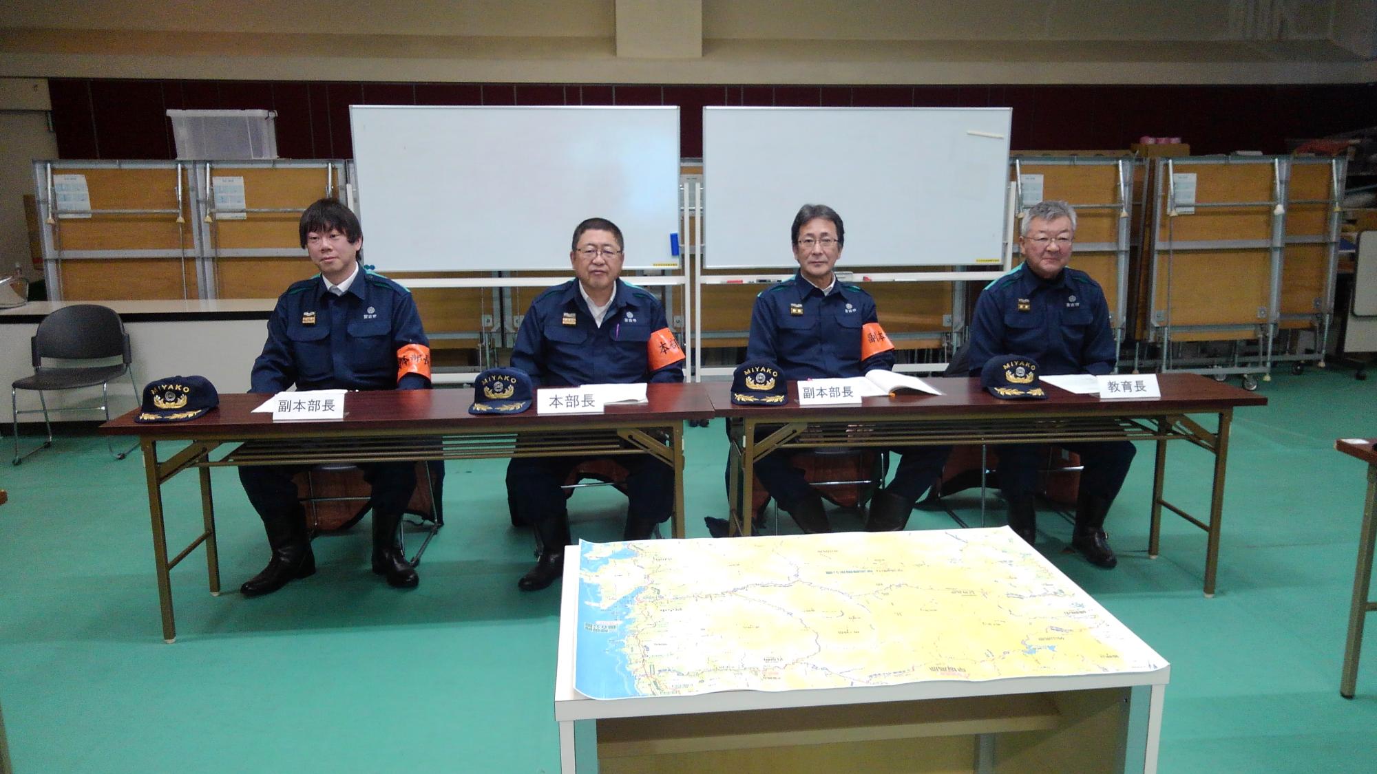 紺色の作業服を着て、左腕にオレンジ色の腕章をつけた市長と3名の関係者の男性が椅子に座っている写真