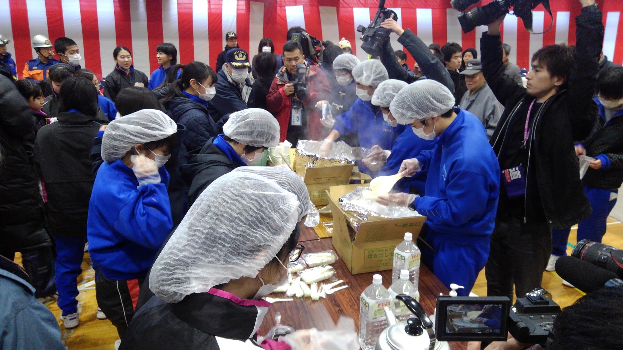 湯気が上がった白ご飯の入った段ボールから、ご飯をよそう様子のヘッドカバーを付けた中学生たちとの写真