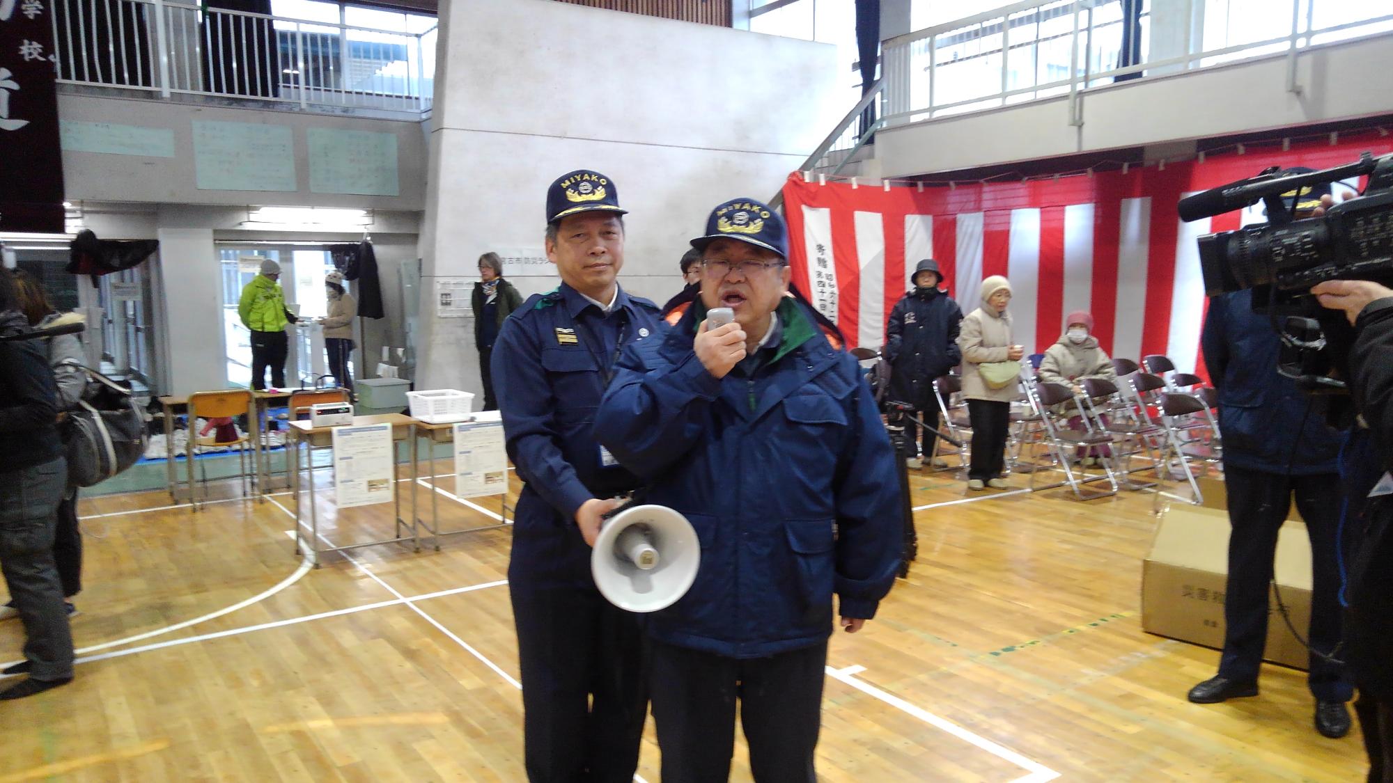 拡声器のマイクで話をする様子の市長と、後方から拡声器を右手で支える様子の男性の写真