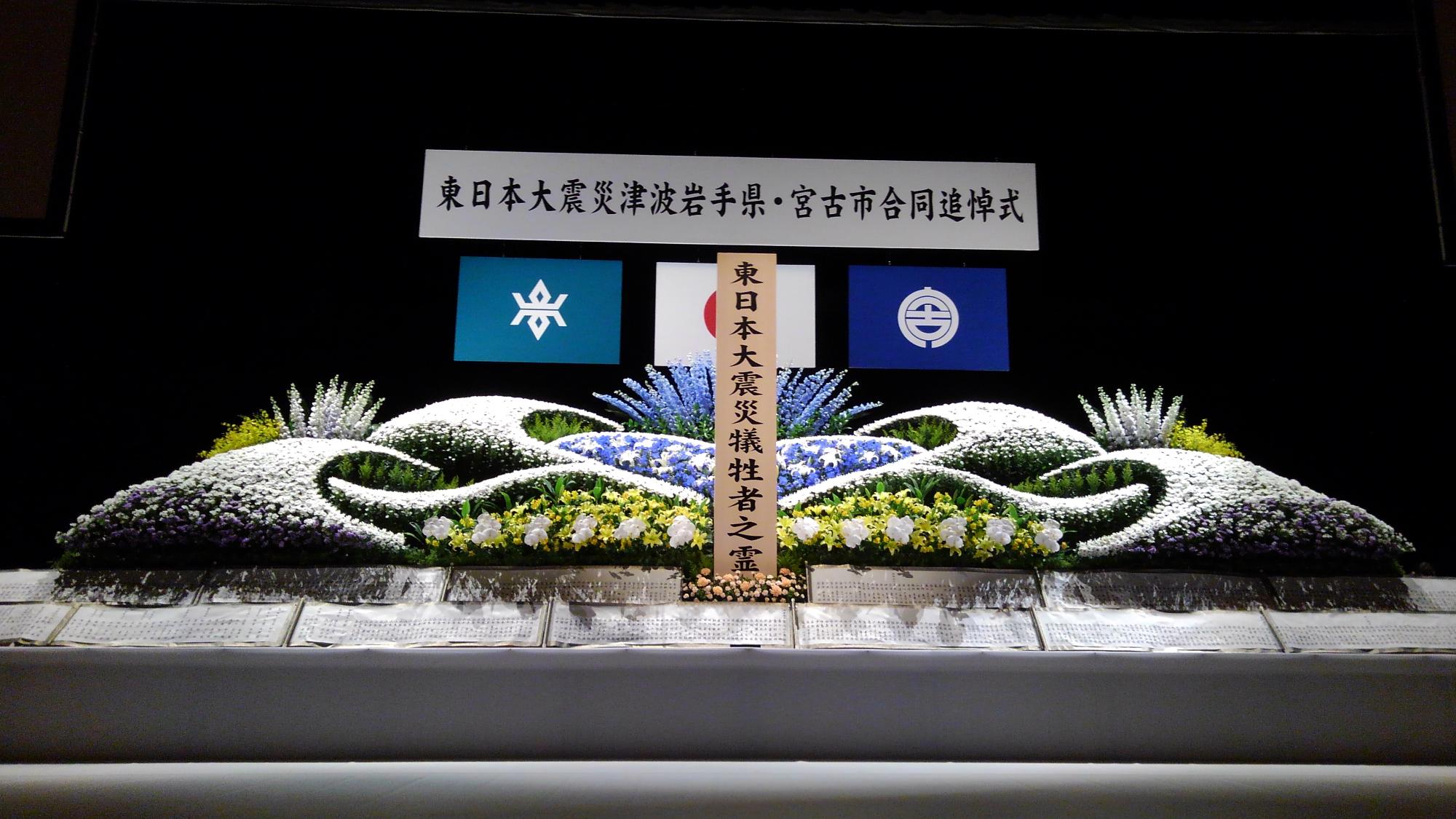 「東日本大震災津波岩手県・宮古市合同追悼式」と書かれた横断幕の下に、白や黄色、青色の花がたくさん設置された献花台の写真