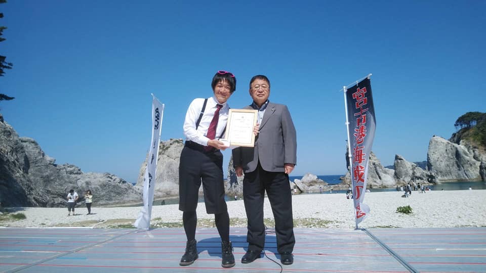 浄土ヶ浜の前で額入りの承認証を持ったデイジーどぶゆきさんと市長が並んでいる記念写真