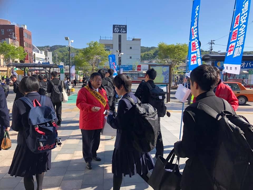 「交通安全運動実施中」と書かれたのぼり旗が立てられた街中で、オレンジ色のジャンパーを着て肩に黄色のタスキを掛けた市長が、登校中の高校生に啓発グッズを手渡し交通安全を呼びかけている様子の写真