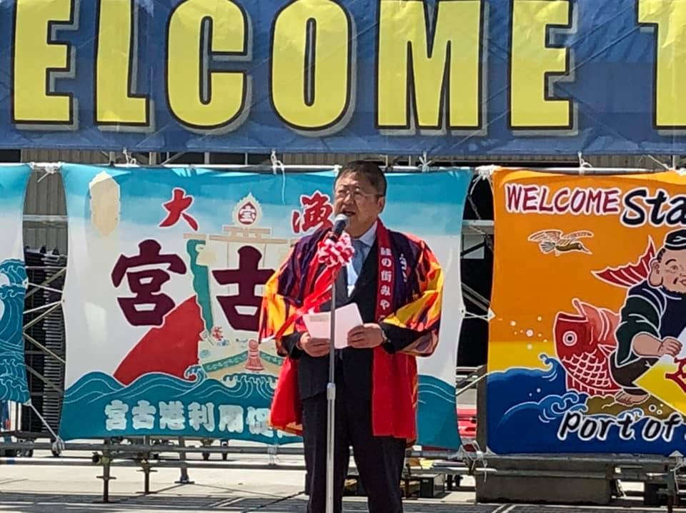 大漁旗が展示された宮古港で、法被を着た市長がマイクスタンドの前で挨拶を行っている写真