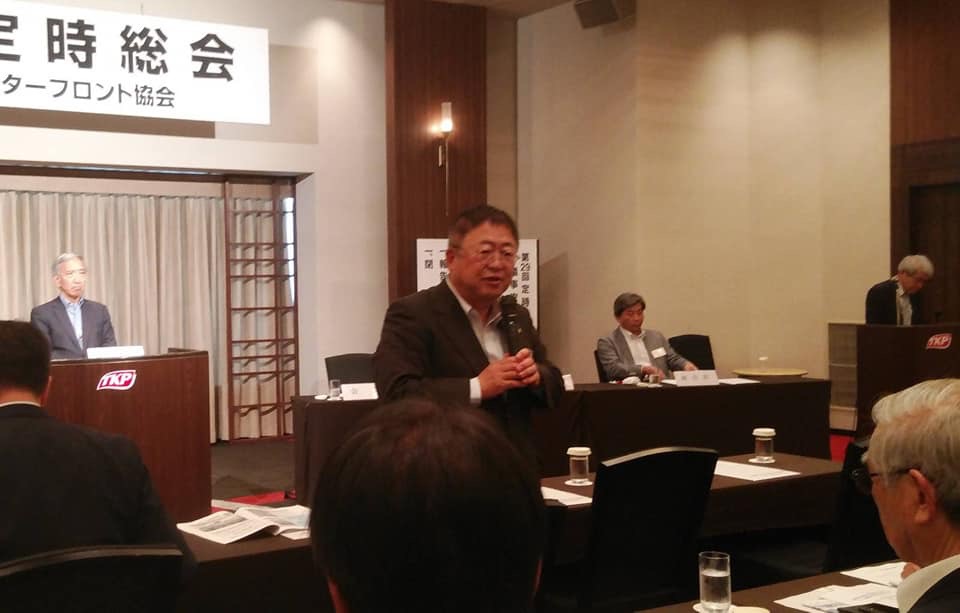 テーブルが並べられ、前方に演台が置かれた会場の中央で、マイクを持ち話をする様子の市長の写真