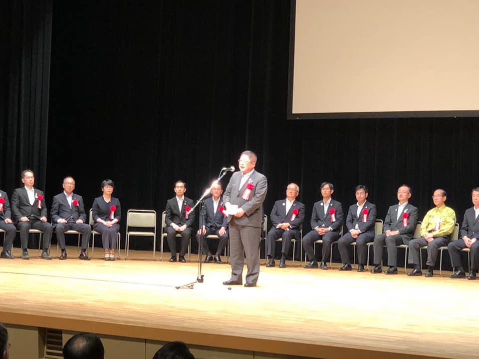 スクリーンのが設置された舞台上で、後方に赤い胸章を着けて並べられた椅子に座る複数の関係者と、舞台の中心に置かれたスタンドマイクに向かい話をする様子の市長の写真