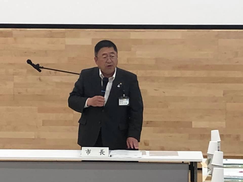 「市長」と書かれたネームプレートが置かれたテーブルの右手を付き、左手にマイクをもって話をする様子の市長の写真