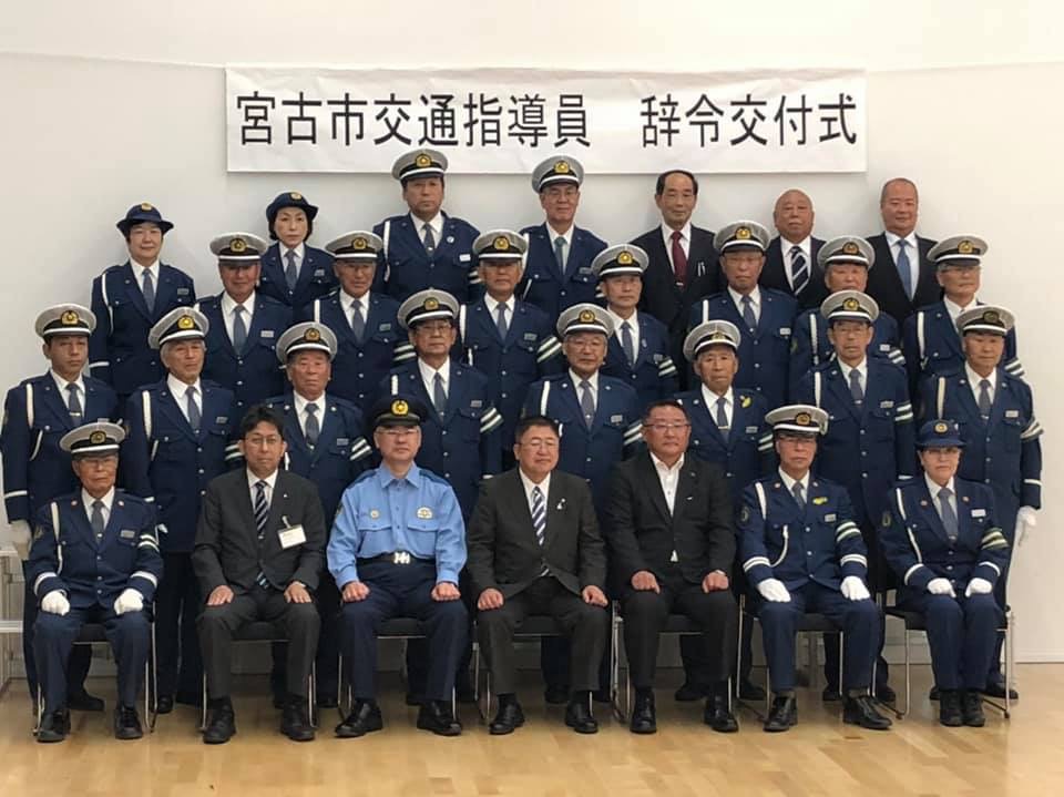 「宮古市交通指導員 辞令交付式」と書かれた横断幕が飾られた壁の前で、1列目は椅子に座った市長と制服を着た指導員やスーツを着た関係者合わせて6名が座り、2列目、3列目、4列目には制帽と制服を着た指導員の方々が立って並んでいる記念写真
