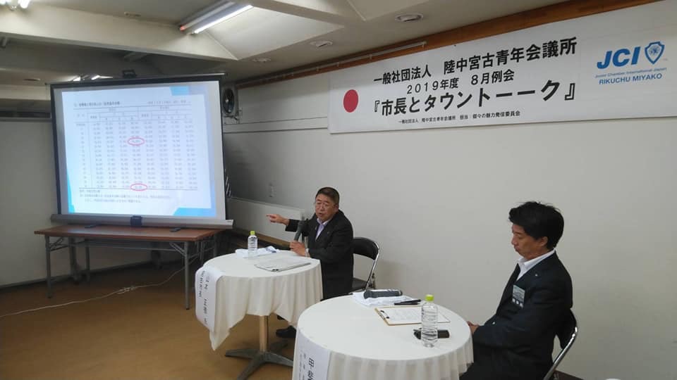 白いテーブルクロスが敷かれた丸テーブルの席に着き、スクリーンを指さす市長と関係者の男性の写真