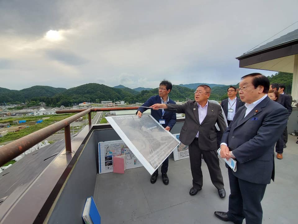 建物の屋上で大きな写真パネルを持つ職員、遠くを指さしながら説明をしている市長、両手をお腹の前に組んで話を聞いている渡辺博道復興大臣の写真