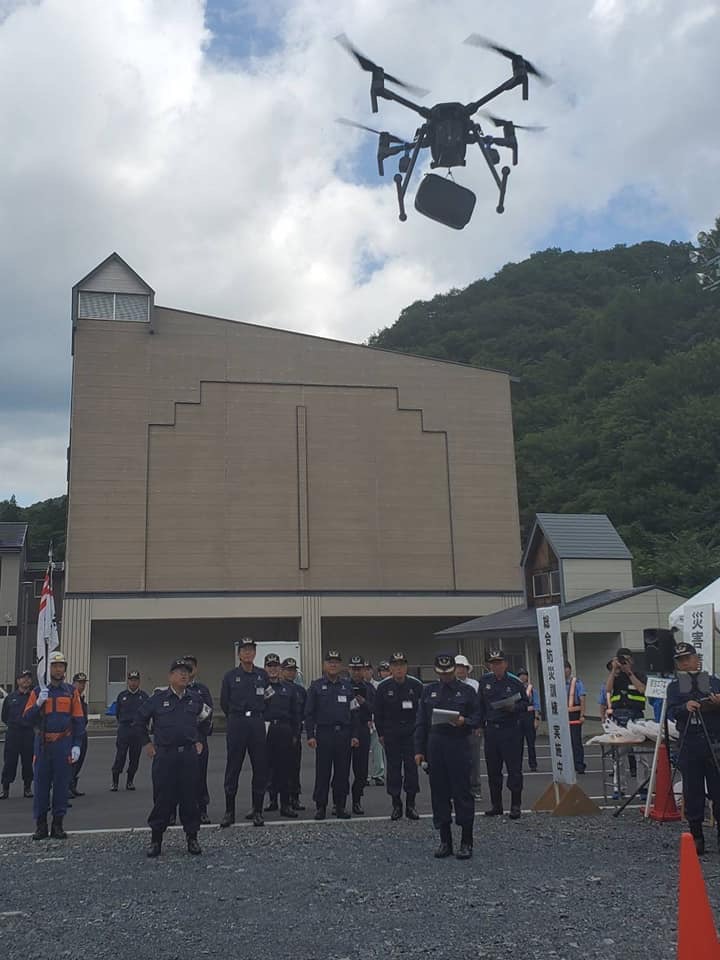 建物の前の広場でドローンが飛んでいる様子を見ている関係者の方々の写真