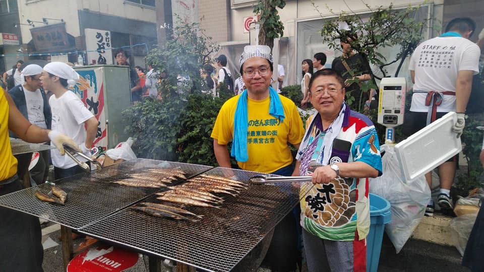 たくさんのサンマが焼かれている大きな網の横に立津市長と、サンマを焼く関係者の方やたくさんのお客さんでにぎわう会場の写真