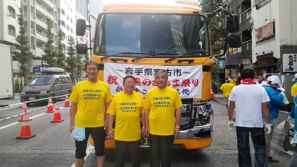 同じ黄色のTシャツを着た市長と男性2人が黄色の大型トラックの前に立っている写真