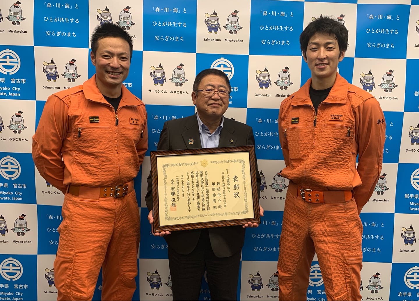 宮古市の会見パネルの前で、表彰状を持つ市長とその両脇に立つオレンジの制服を着た消防隊員の方の写真