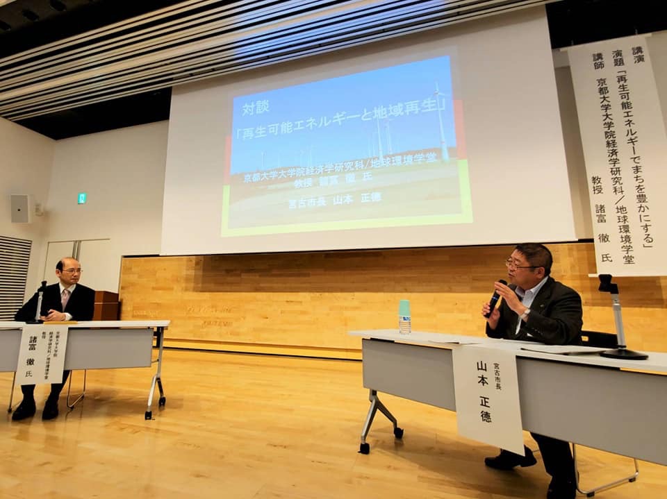 題目が投影されたスクリーンの前にハの字に置かれた長机に市長と諸富教授が座り、対談をしている様子の写真