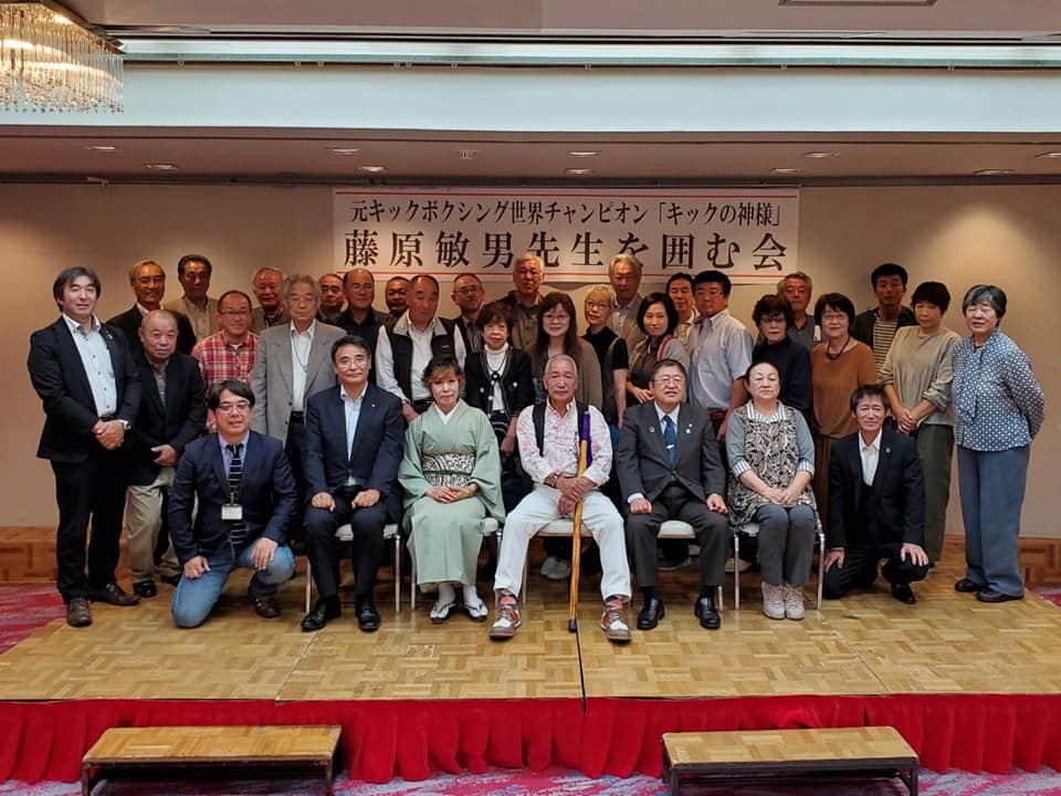 藤原敏男先生を囲む会と書かれた横断幕を背景に、前列中央に藤原敏男さんが座り、市長含むその他30名程の関係者の方々が3列に並んだ集合写真