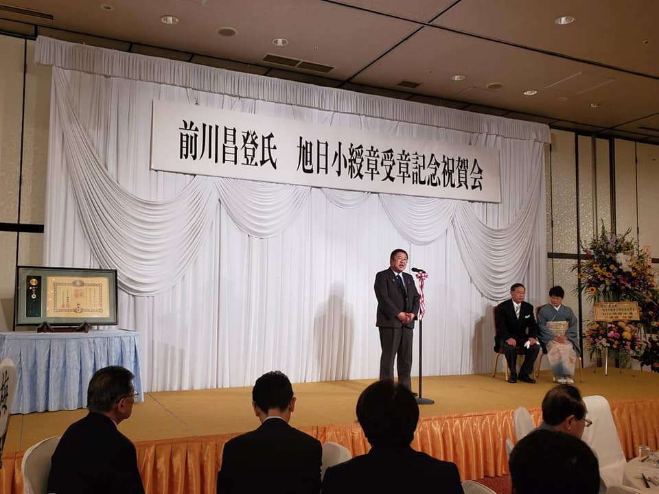 前川昌登氏 旭日小綬章受章記念祝賀会と書かれた横断幕がはられたステージ上で、マイクの前に立ち挨拶をする市長の写真