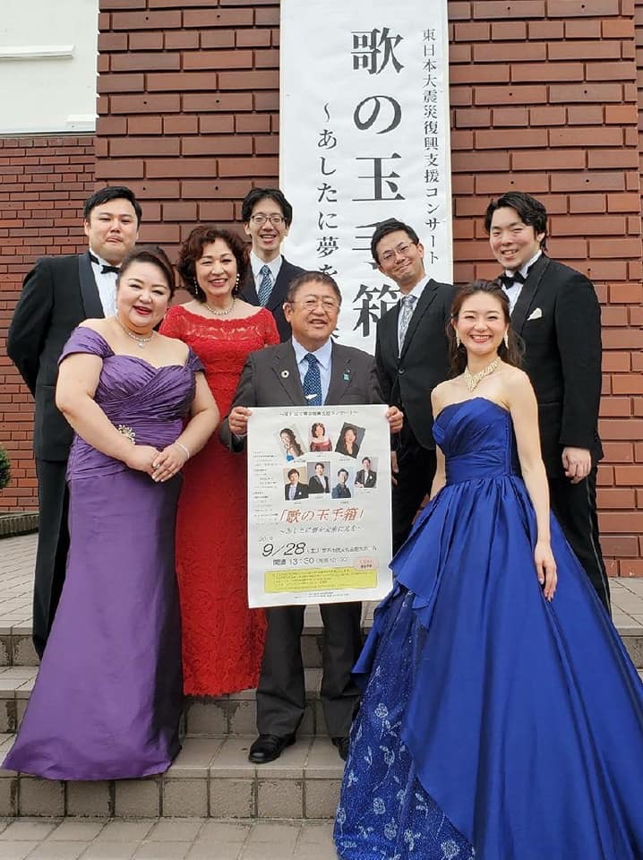 会場外の立て看板の前で歌の玉手箱のポスターを持った市長と、ドレスやタキシードを着た出演者男女7名の集合写真