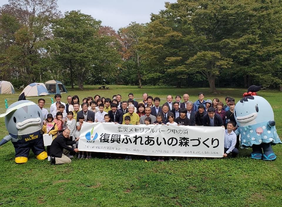 テントも張られている自然豊かな公園の中で撮影された活動報告会に参加した多くの関係者や子供たちと、宮古市のゆるキャラサーモンくん・みやこちゃんの集合写真