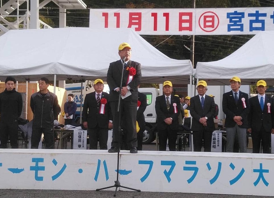 台の上で黄色い帽子を被りスタンドマイクに向かい話をする様子の市長の写真