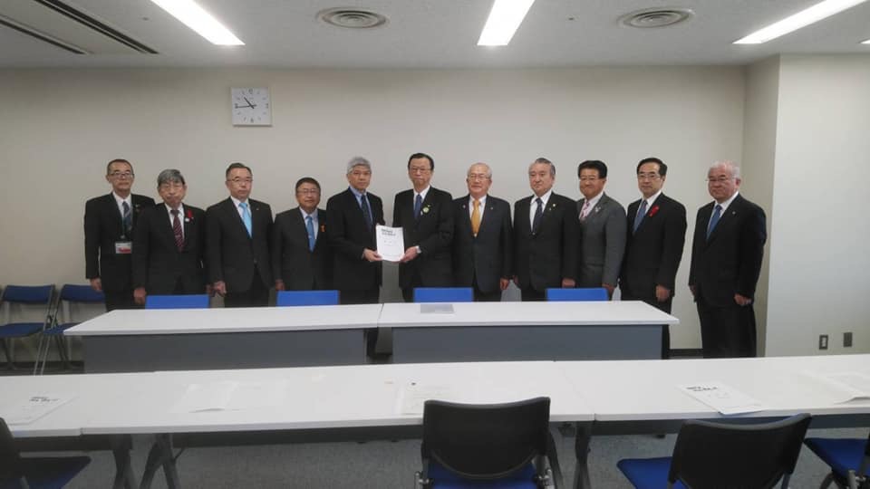 テーブルが並んだ部屋の壁際で、要望書を一緒に持つ2名の男性を中心に、市長と8名の関係者の男性が並んでいる記念写真