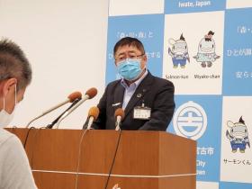 臨時記者会見の会場の後方に宮古市のバックパネルがあり演台の上に数個設置されたスタンドマイクの前で市長がマスコミに向けて報告している写真