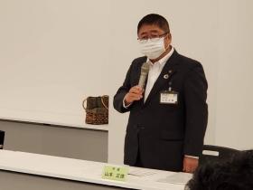 委員会の会場で市長がネームプレートがテーブルに置かれた自席に立ち、男女共同参画実施計画や男女共生センターの実績について述べている写真