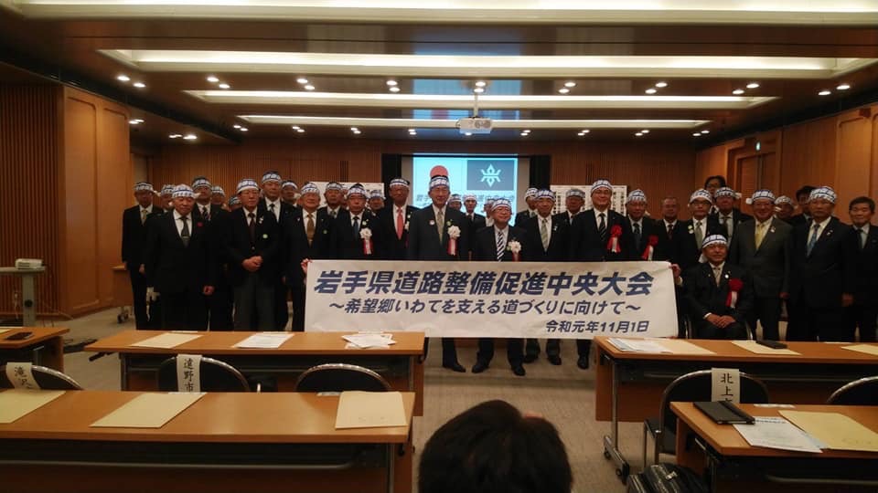 「岩手県道路整備促進中央大会」と書かれた横断幕の後方で、頭に鉢巻を巻き胸章を着けた関係者の方々が立って並び記念撮影をしている写真