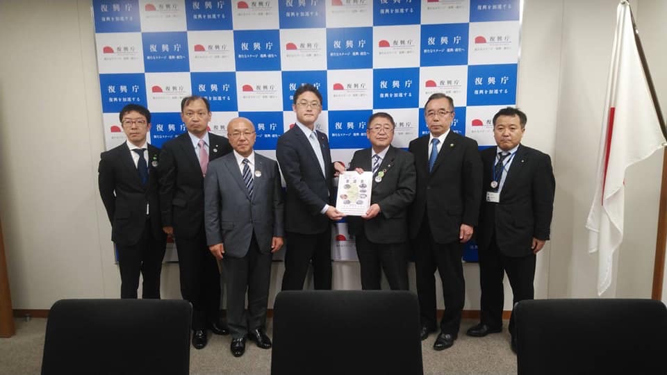 バックパネルの前で要望書を一緒に持つ市長と男性1名、関係者の男性5名が横一列に並んでいる記念写真
