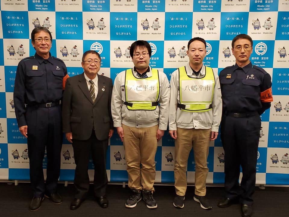 バックパネルの前で作業服に黄色いゼッケンベストを身に着けた男性が2名、スーツを着た市長、紺色の作業着を着た男性2名が横一列に並んでいる記念写真
