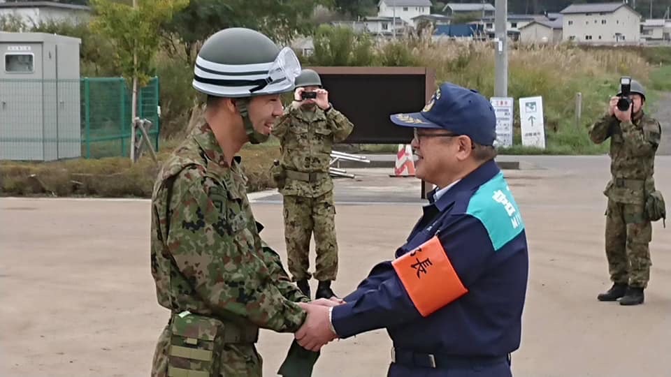 紺色の作業服にオレンジ色の腕章をつけた市長が、迷彩服を着た自衛隊員と両手で握手をしている様子の写真