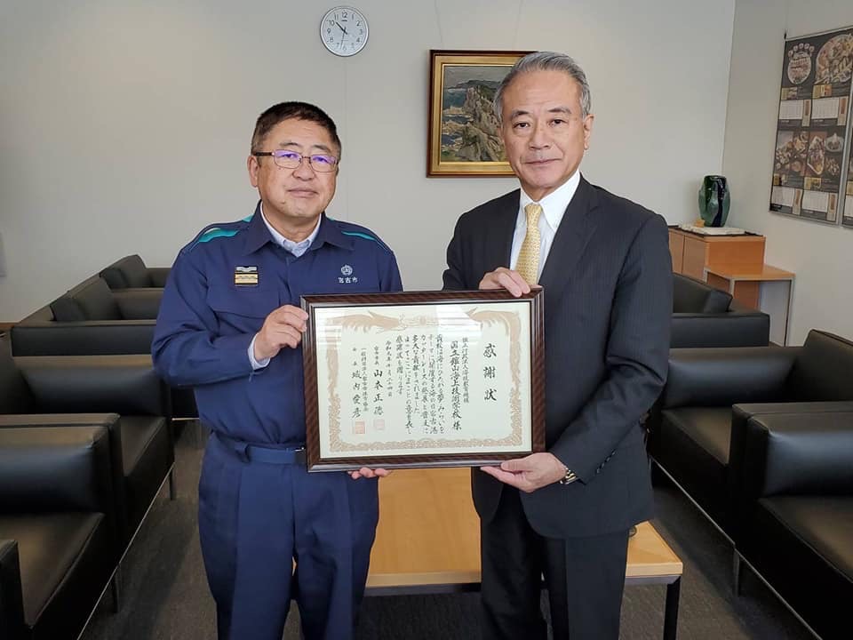 紺色の作業服を着た市長と野崎哲一理事長が一緒に感謝状の額縁を持って記念撮影をしている写真