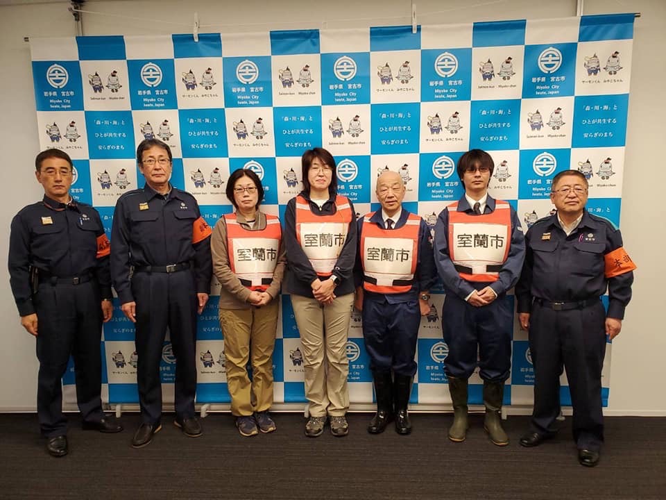 バックパネルの前で、「室蘭市」と書かれたオレンジ色のゼッケンベストを身に着けている4名の方々と、紺色の作業服を着た市長と2名の男性が横一列に並んでいる記念写真