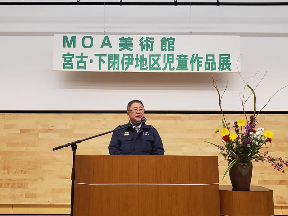 「MOA美術館 宮古・下閉伊地区児童作品展」と書かれた横断幕が吊るされた舞台上で、演台の前で話をする様子の市長の写真
