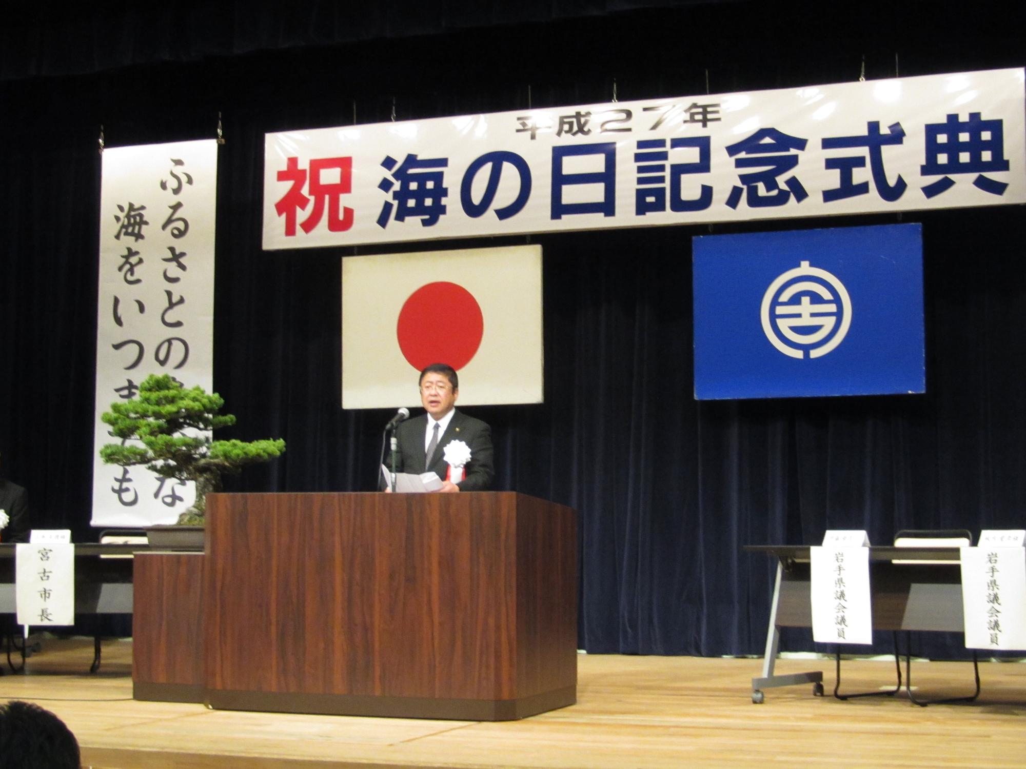 「平成27祝海の日記念式典」と書かれた横断幕、日本国旗、宮古市の市町村旗が掲げられた舞台で、演台に置かれたスタンドマイクに向かい話をする市長の写真