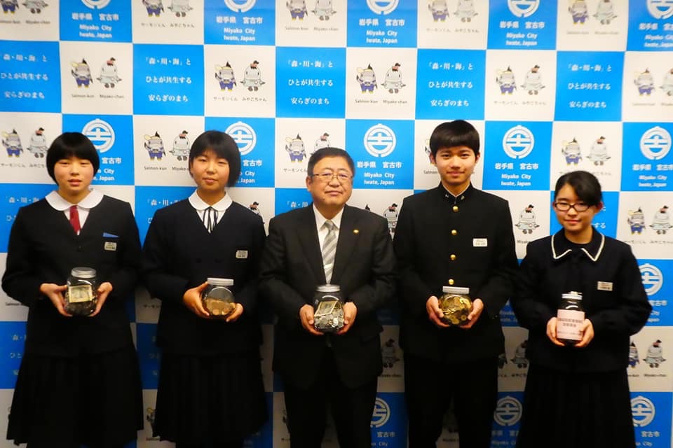 バックパネルの前で募金の入った瓶を持つ市長と制服を着た中学生4名の記念写真