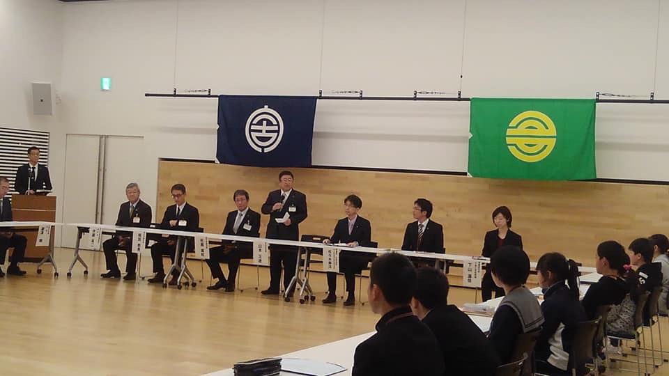 コの字に並べられたテーブルの席に着く関係者の方々や小中学生たちと、右手にマイクを持ち立って話す様子の市長の写真