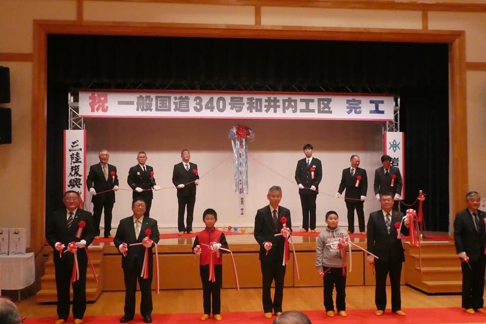 「祝 一般国道340号和井内工区 完工」と書かれた横断幕とくす玉が吊るされた舞台に6名の男性が立ち、舞台の下でテープカットをする5名の男性と男の子2名を写した写真
