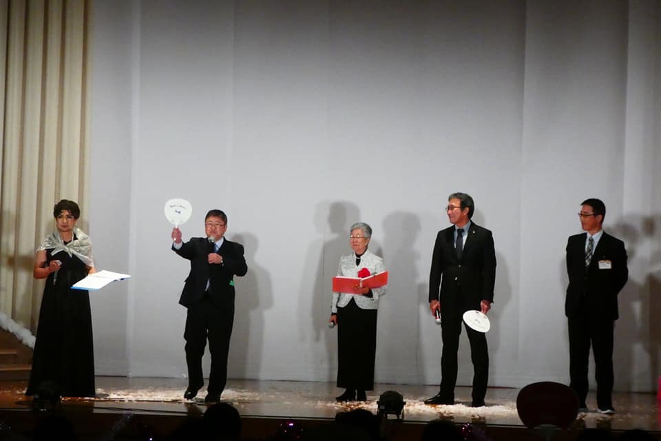 台本を右手に持つ2名の女性、右手で持ったうちわを頭上に掲げる市長、関係者の男性2名が横一列に並んでいる写真