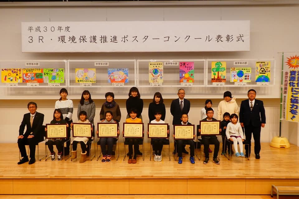 「平成30年度3R・環境保護推進ポスターコンクール表彰式」と書かれた横断幕の下に、ポスターが展示された舞台上で、前列で賞状を持ち椅子に座る7名の子供たちと白い上着を着た1名の子供、関係者の男性、2列目に子供2名と市長を含む9名の方々が立って並んでいる記念写真